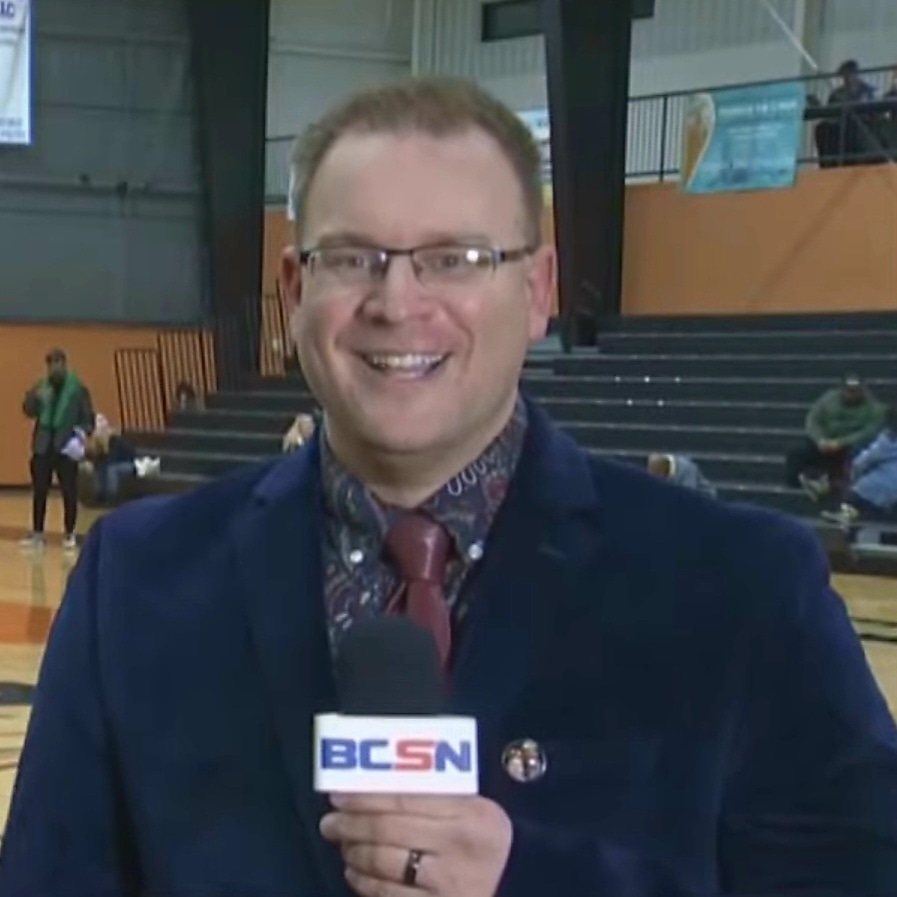 Tim Clagg smiling on camera with a BCSN microphone during a live gymnasium broadcast
