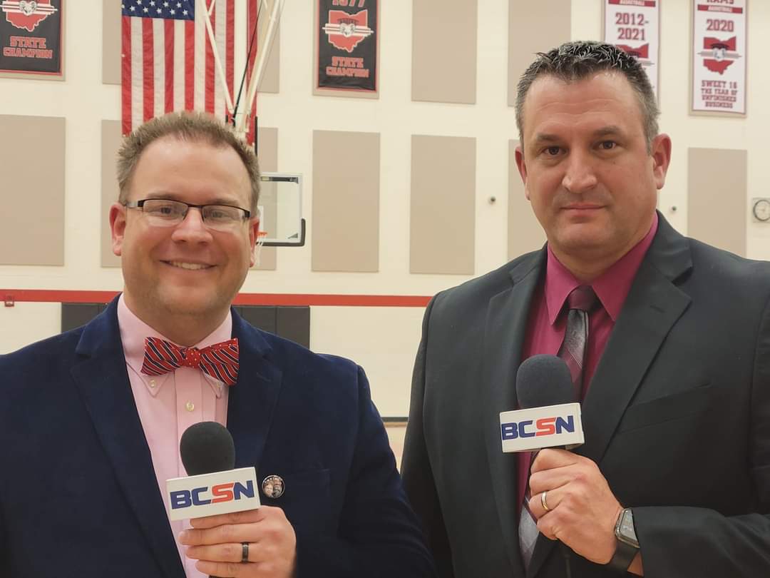 Tim Clagg in a bow tie alongside his BCSN analyst inside a championship-banner gymnasium